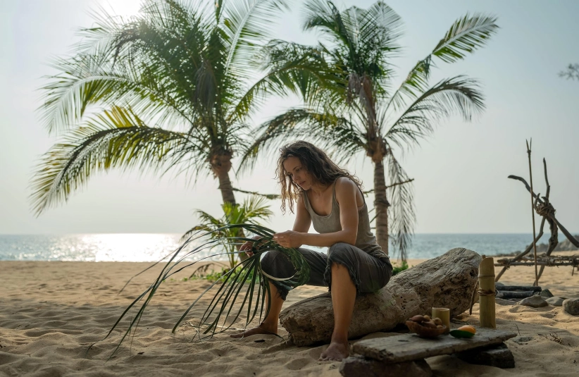 Rachel McAdams in Send Help (2026) sits on a large driftwood log on a sandy beach, weaving palm fronds. Palm trees and the ocean are in the background, creating a serene scene.