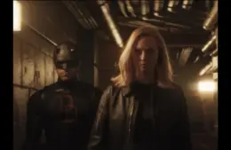 Daredevil stands in a dimly lit corridor, accompanied by Karen with blonde hair in a black leather jacket, conveying a tense, mysterious Daredevil: Born Again scene