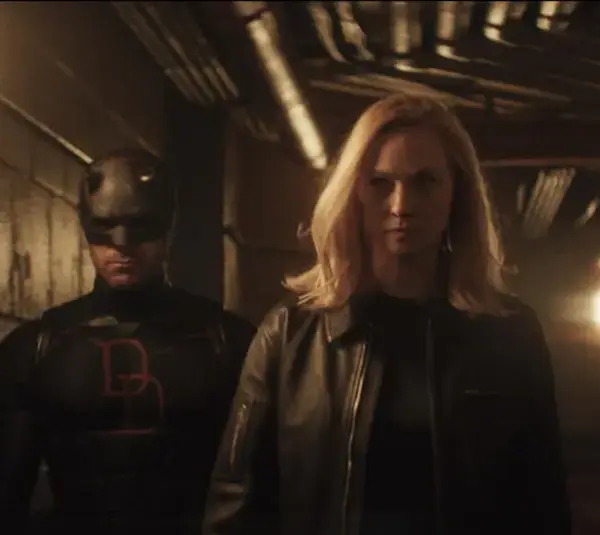 Daredevil stands in a dimly lit corridor, accompanied by Karen with blonde hair in a black leather jacket, conveying a tense, mysterious Daredevil: Born Again scene