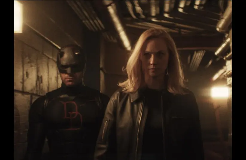 Daredevil stands in a dimly lit corridor, accompanied by Karen with blonde hair in a black leather jacket, conveying a tense, mysterious Daredevil: Born Again scene