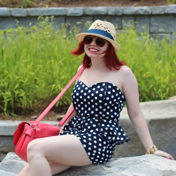 Sweetheart neckline dotted playsuit styled with a red bag, aviators, and a hat trimmed with a navy‑blue ribbon, showcasing chic polka dots effortlessly.