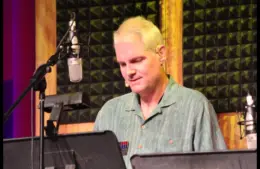 Tom Kane, a person with short blond hair is speaking into a microphone in a recording studio. They wear a green shirt and have a focused expression.