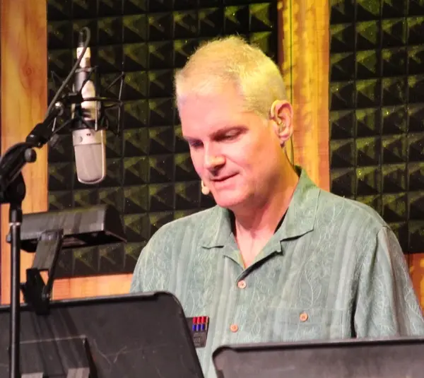 Tom Kane, a person with short blond hair is speaking into a microphone in a recording studio. They wear a green shirt and have a focused expression.