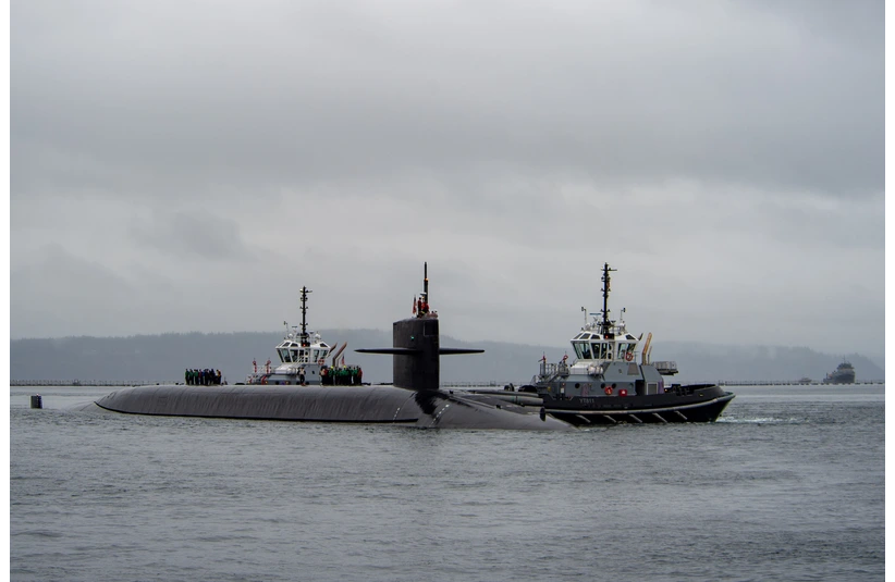 A gray submarine navigates a calm, cloudy sea, accompanied by two tugboats. The scene is serene and industrial, emphasizing maritime engineering, USS Nebraska