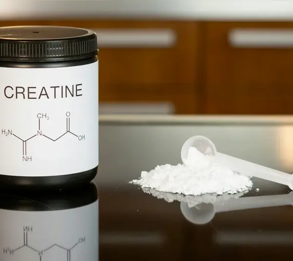 A black container labeled "Creatine" with a chemical formula sits on a reflective surface. Beside it, a scoop rests on a pile of white powder.