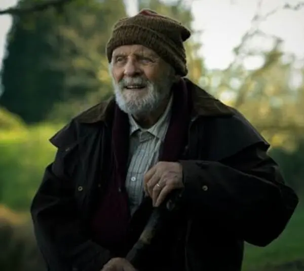 Anthony Hopkins from A Visit To Grandpa's, with a gentle smile, stands amidst lush greenery. He wears a brown jacket, a beanie, and holds a walking stick, exuding warmth and serenity.