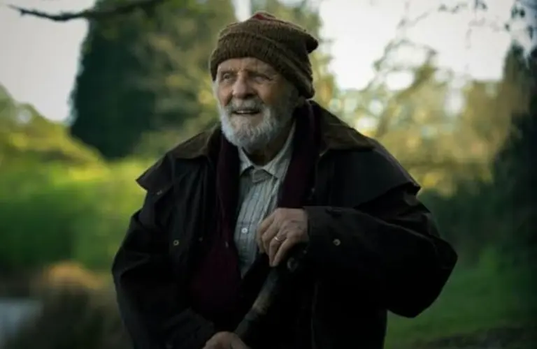 Anthony Hopkins from A Visit To Grandpa's, with a gentle smile, stands amidst lush greenery. He wears a brown jacket, a beanie, and holds a walking stick, exuding warmth and serenity.