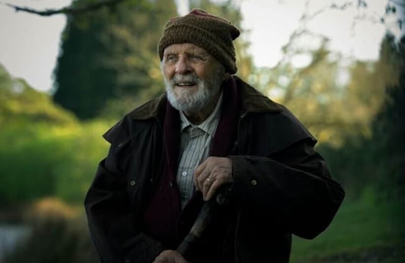 Anthony Hopkins from A Visit To Grandpa's, with a gentle smile, stands amidst lush greenery. He wears a brown jacket, a beanie, and holds a walking stick, exuding warmth and serenity.