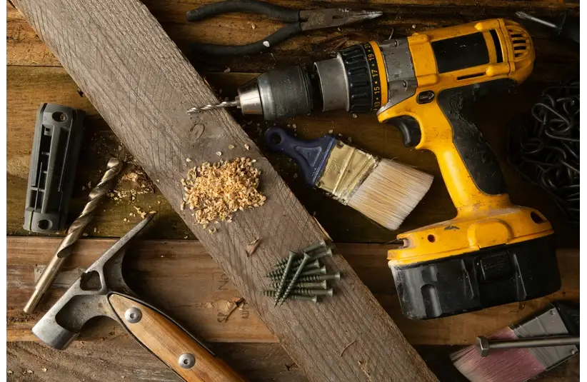 A yellow cordless drill, hammer, drill bits, screws, and paintbrush are scattered on a wooden surface, conveying a workshop or DIY setting.