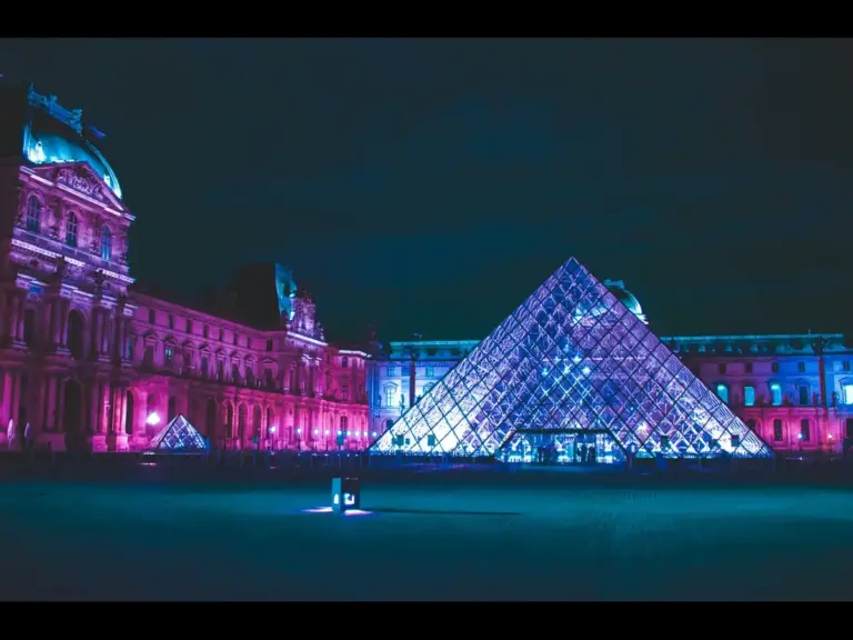 Illuminated glass pyramids at the Louvre Museum contrast with historic architecture at night. The scene glows in vibrant blue and pink lights. Museum and noctourism.