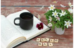 An open book with a coffee cup and some spring flowers with the words "Hello May" written in blocks. What will the book of the month be for May?