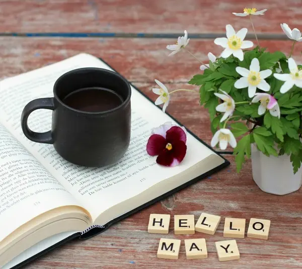 An open book with a coffee cup and some spring flowers with the words "Hello May" written in blocks. What will the book of the month be for May?