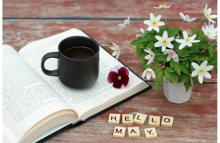 An open book with a coffee cup and some spring flowers with the words "Hello May" written in blocks. What will the book of the month be for May?