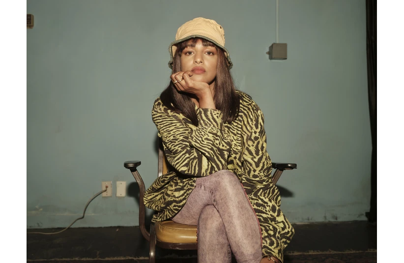 M.I.A in a patterned shirt and beige hat sits on a chair against a plain wall. They rest their chin on their hand, appearing thoughtful.