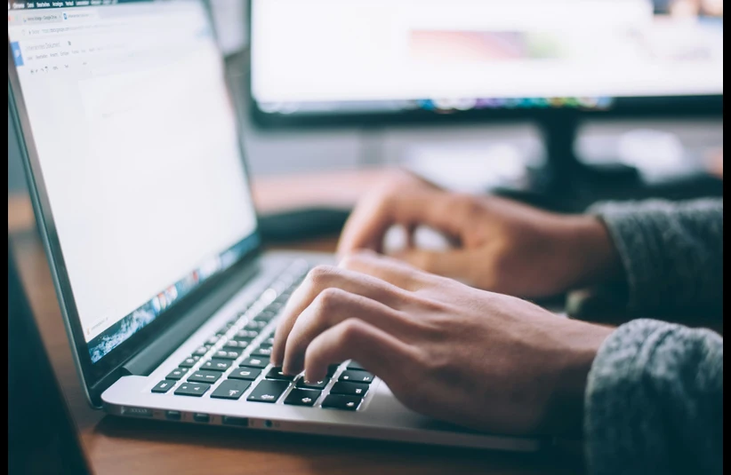 Hands typing on a laptop keyboard, with a blurred monitor in the background. The scene conveys focus and productivity in a workspace.