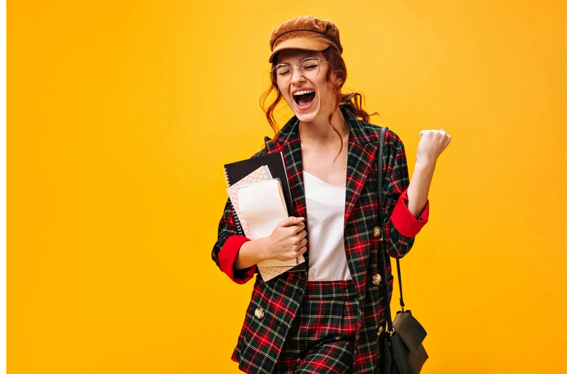 Joyful woman in a plaid suit and cap, holding notebooks, fist raised in triumph against a bright yellow background. She exudes excitement and confidence.