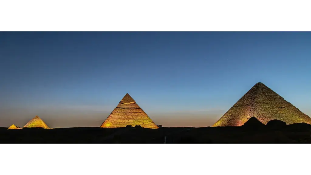 Three pyramids illuminated against a twilight sky; the scene conveys a sense of ancient majesty and calm with deep blues and warm hues. Excellent time for noctourism.