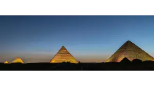 Three pyramids illuminated against a twilight sky; the scene conveys a sense of ancient majesty and calm with deep blues and warm hues. Excellent time for noctourism.