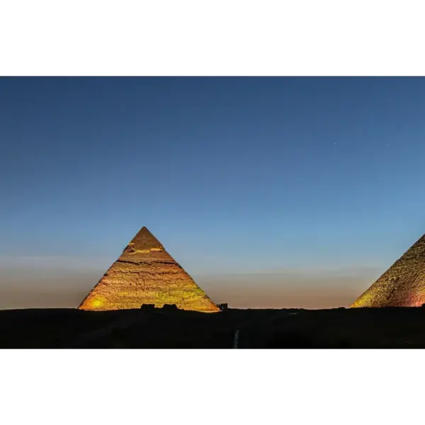 Three pyramids illuminated against a twilight sky; the scene conveys a sense of ancient majesty and calm with deep blues and warm hues. Excellent time for noctourism.