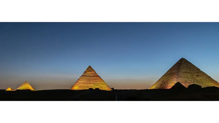 Three pyramids illuminated against a twilight sky; the scene conveys a sense of ancient majesty and calm with deep blues and warm hues. Excellent time for noctourism.