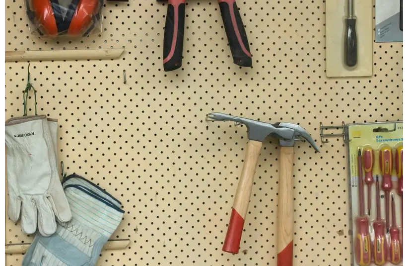 A pegboard displays various tools: a pair of work gloves, ear protection, a hammer, pliers, a screwdriver, and a set of packaged screwdrivers. Neatly organized workspace.
