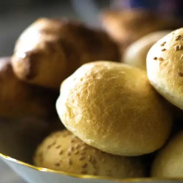 A bowl filled with various rolls and buns placed on a wooden table. Refined carbs, bread