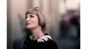 Stylish portrait of a woman with a blonde soft bob haircut and sunglasses, looking up with a blurred urban background.