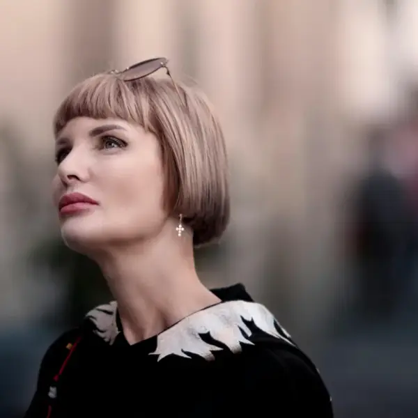 Stylish portrait of a woman with a blonde soft bob haircut and sunglasses, looking up with a blurred urban background.
