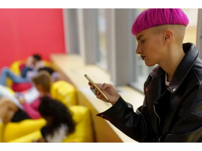 A stylish woman with pink hair uses her smartphone while sitting indoors on bright bean bags, embracing vibrant color drenching energy.