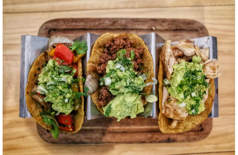 Three vibrant tacos with fresh guacamole, veggies, and meat on a wooden tray perfect for budget dates