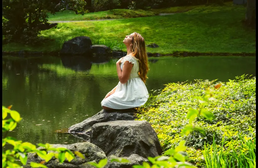 In daily devotional, a woman sits by a lake when hand over her heart meditating on God's goodness.