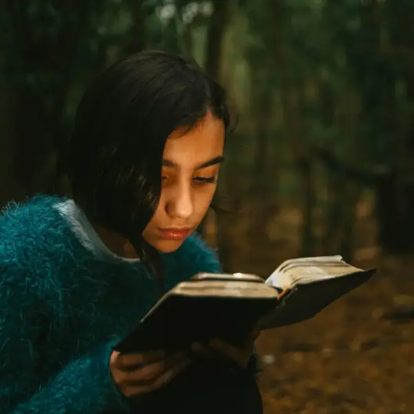 In daily devotional, a young woman reads a Bible outside surrounded surrounded by trees.