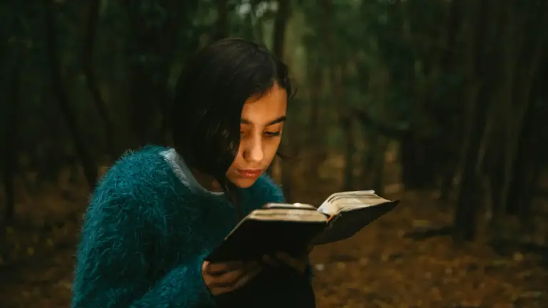 In daily devotional, a young woman reads a Bible outside surrounded surrounded by trees.