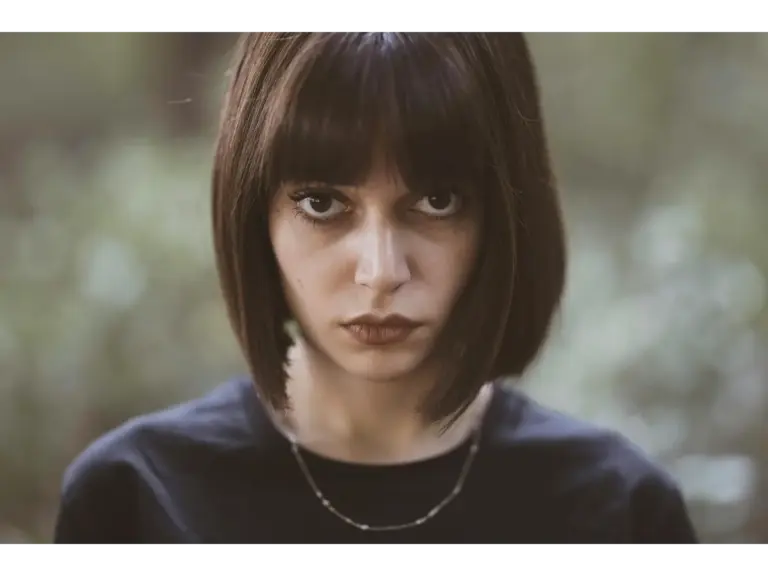 Close-up portrait of a serious woman with a short dark soft bob and intense gaze, outdoors.