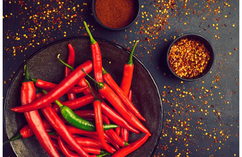 Close-up of fresh red and green chili peppers in a bowl with spices and pepper flakes, highlighting the ingredients driving the spicy craze.