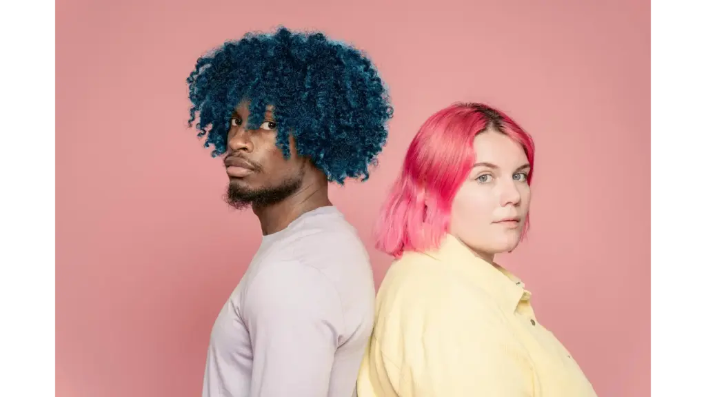 A young diverse couple with blue and pink hair posing back to back against a pink background, showcasing bold color drenching vibes.