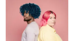A young diverse couple with blue and pink hair posing back to back against a pink background, showcasing bold color drenching vibes.
