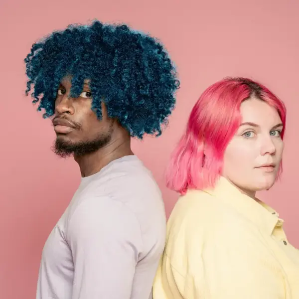 A young diverse couple with blue and pink hair posing back to back against a pink background, showcasing bold color drenching vibes.