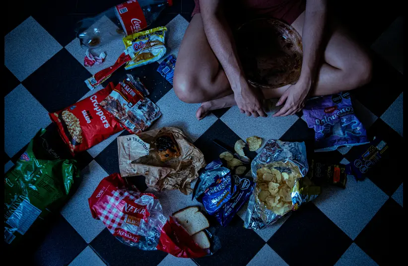 Person sitting on a checkered floor surrounded by snacks, chips, bread, and empty packages. The scene conveys a sense of messiness and indulgence.