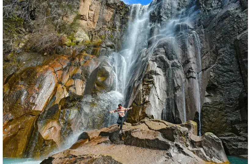 A person poses on rocks in front of a waterfall