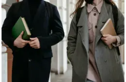 Two students carrying books and wearing coats while walking through a building hallway dressed in academic style