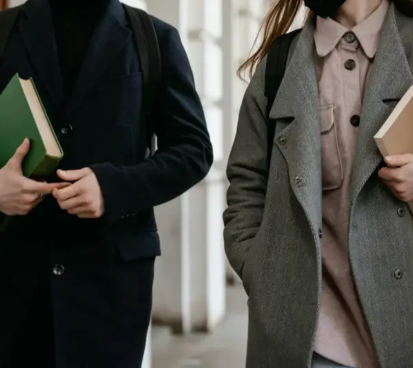 Two students carrying books and wearing coats while walking through a building hallway dressed in academic style