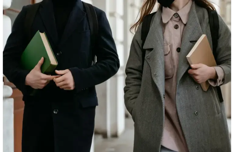 Two students carrying books and wearing coats while walking through a building hallway dressed in academic style