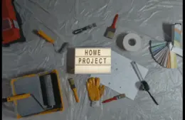 Tools and supplies for a home project are laid out on a plastic-covered surface. Items include paint rollers, brushes, a measuring tape, color samples, and gloves. A light box in the center displays "HOME PROJECT," conveying a DIY, home workshop theme.