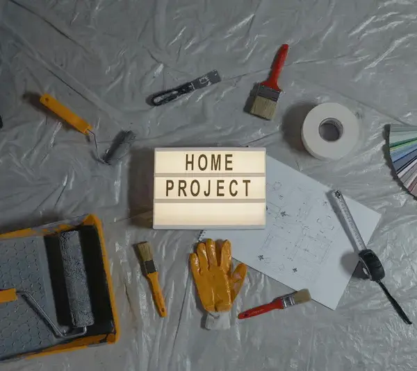 Tools and supplies for a home project are laid out on a plastic-covered surface. Items include paint rollers, brushes, a measuring tape, color samples, and gloves. A light box in the center displays "HOME PROJECT," conveying a DIY, home workshop theme.