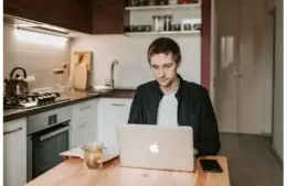 A young man using a laptop in a cozy kitchen, illustrating remote work lifestyle.