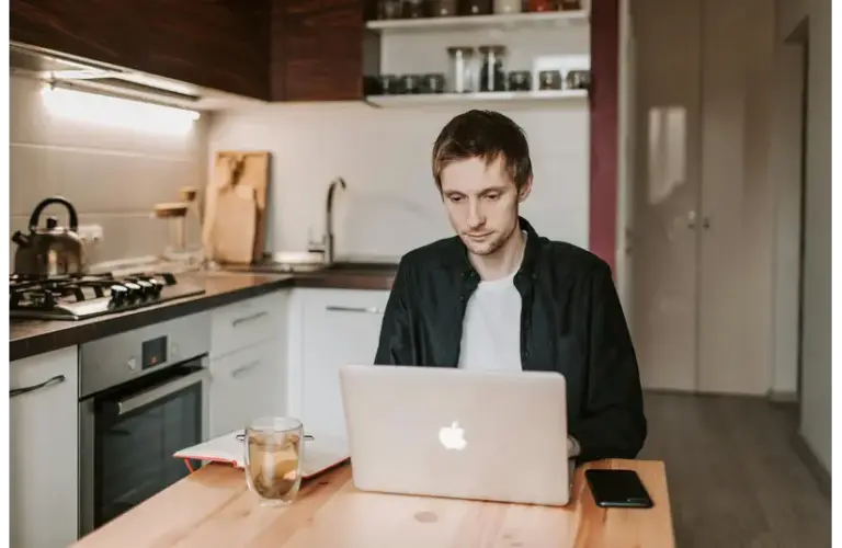 A young man using a laptop in a cozy kitchen, illustrating remote work lifestyle.
