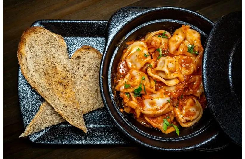A black plate holds toasted bread and a bowl of ravioli in tomato sauce, garnished with herbs, creating a warm and inviting presentation.