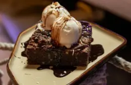 A rich chocolate brownie topped with two scoops of vanilla ice cream, drizzled with chocolate sauce. Served on a beige plate, exuding a tempting, indulgent vibe, dessert, date night