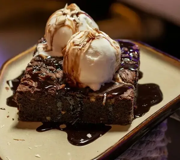 A rich chocolate brownie topped with two scoops of vanilla ice cream, drizzled with chocolate sauce. Served on a beige plate, exuding a tempting, indulgent vibe, dessert, date night